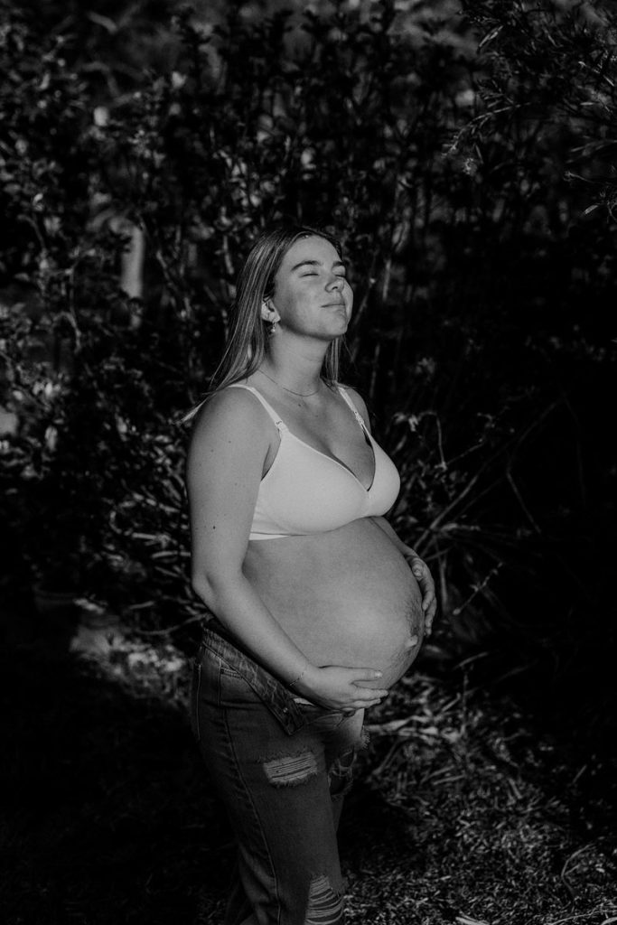 black and white image of a young pregnant lady facing the sun  and holding her belly
