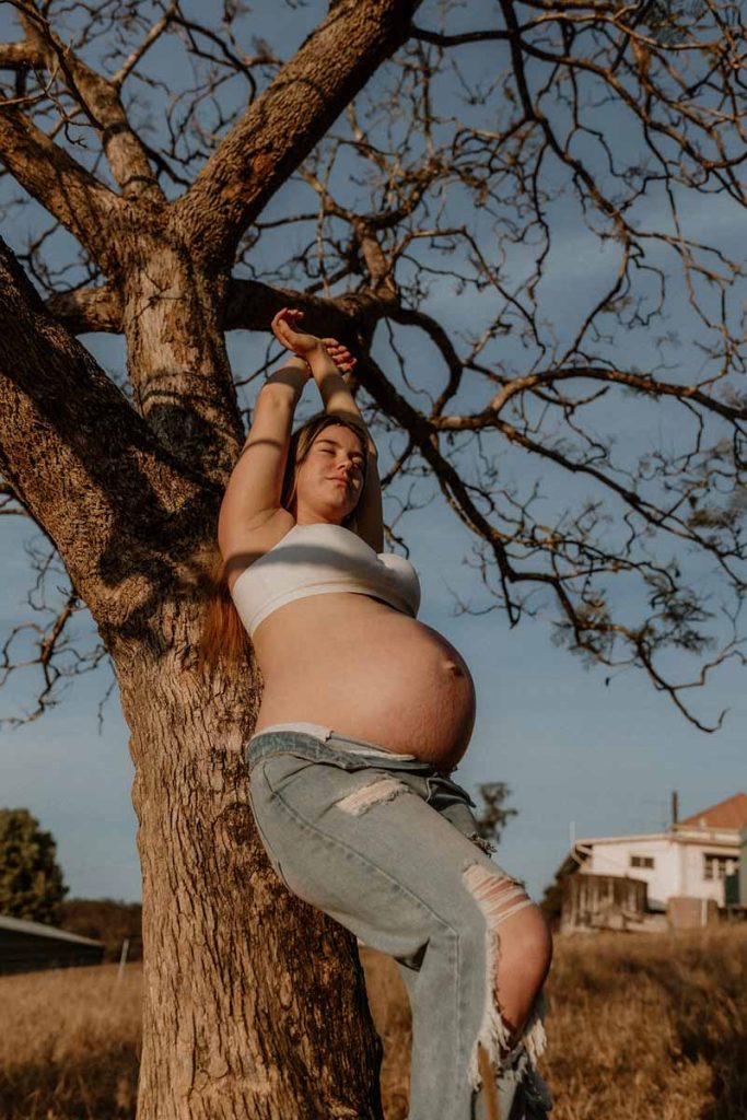 young pregnant woman leaning on a tree with her arms up