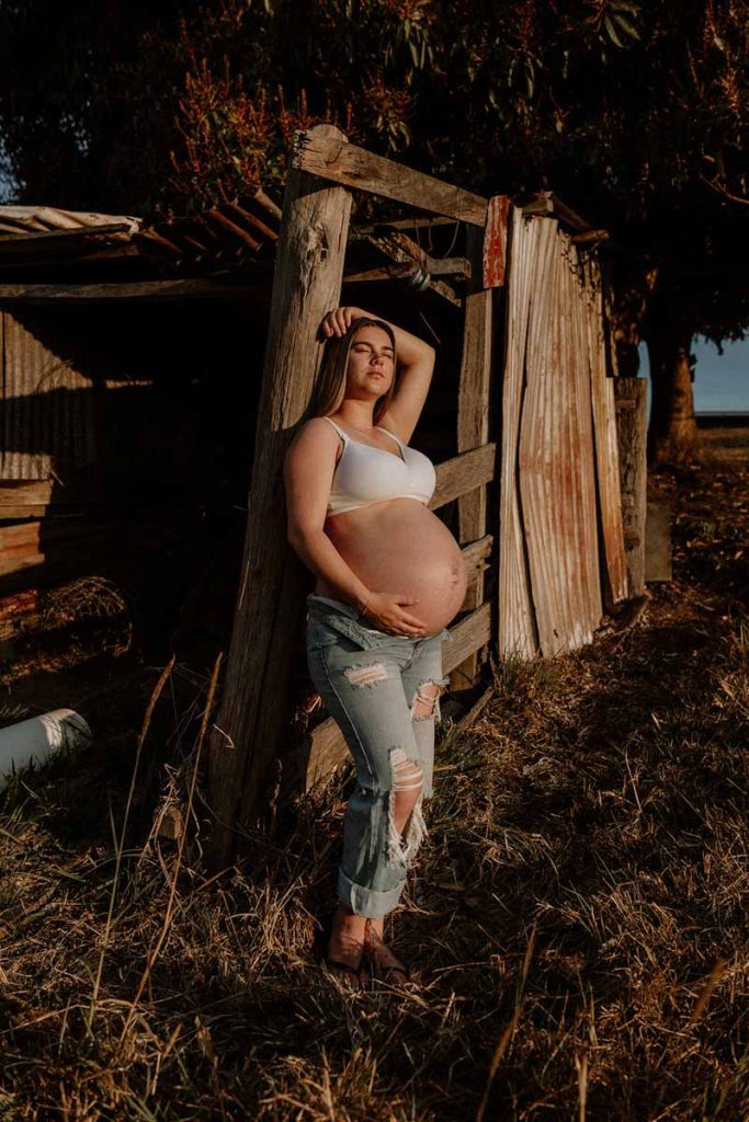 young pregnant leaning on a wooden barn looking straight 