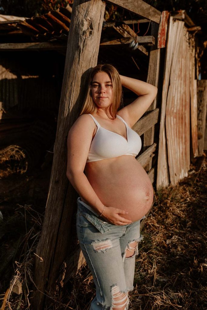young pregnant leaning on a wooden barn looking straight 