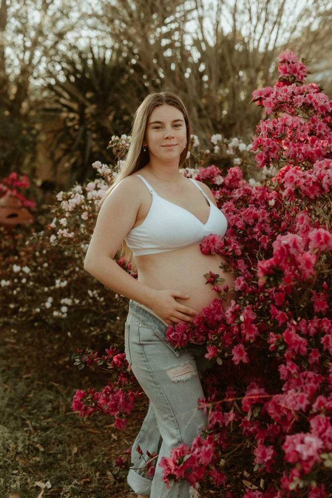 young pregnant lady standing in front of a pink flowers bushes