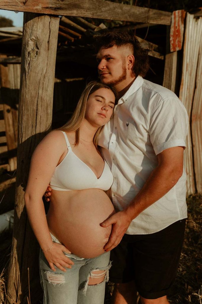pregnant young couple lining on a barn she has a eyes closed and his holding her belly