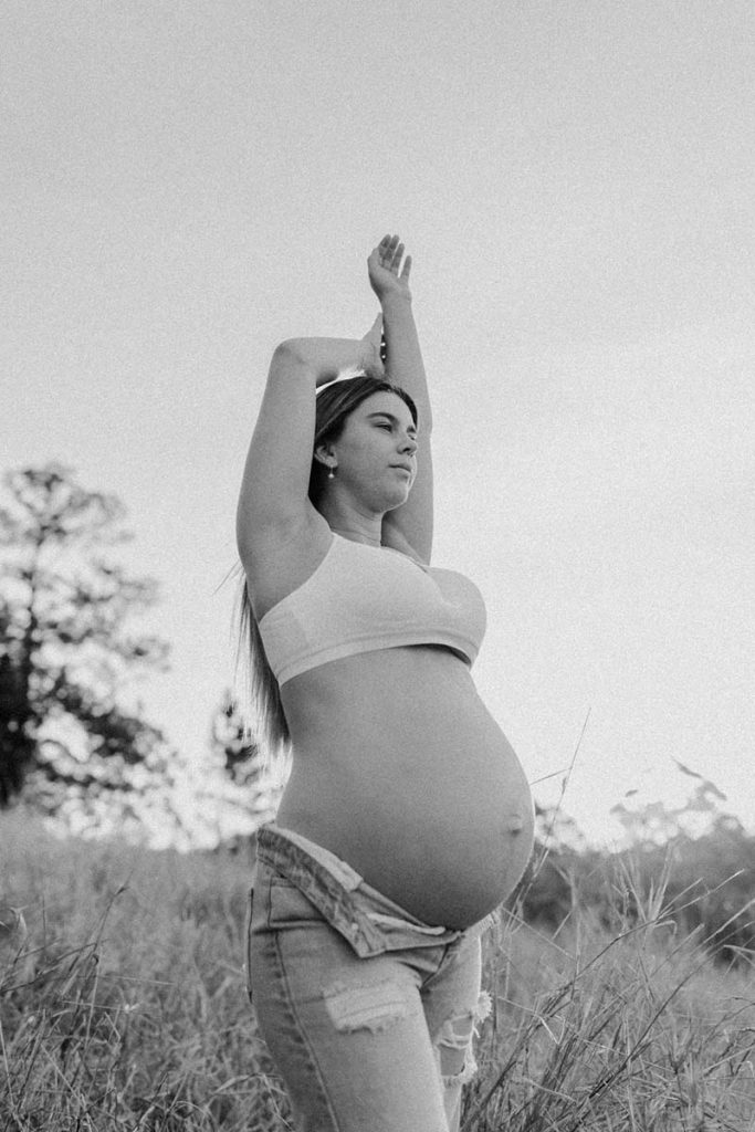 young pregnant woman in a field with long grass arms up and looking staright