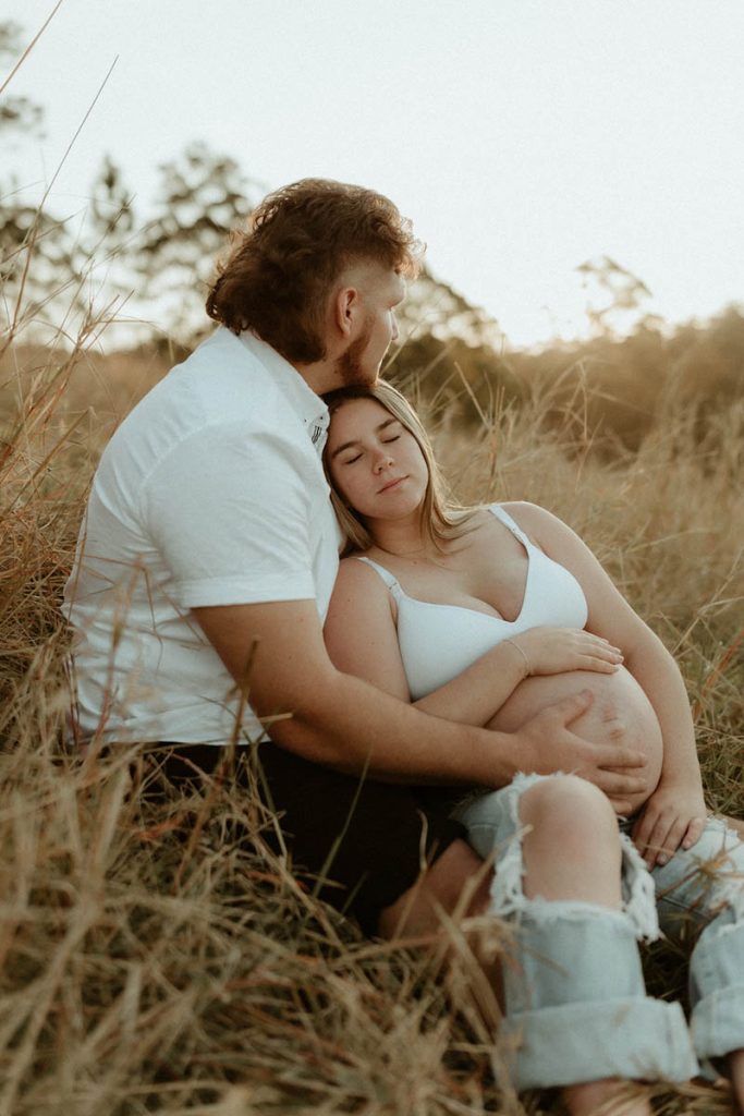pregnant couple snuggling sitting in a long grass the woman has a eyes closed