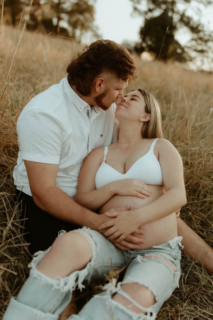 Pregnant couple sitting in the long grass kissing both their hand on the bump