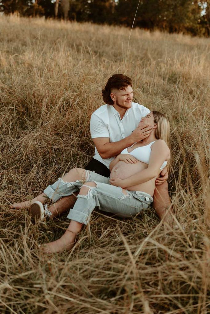 pregnant couple sitting in the long grass snuggling and smiling at each other