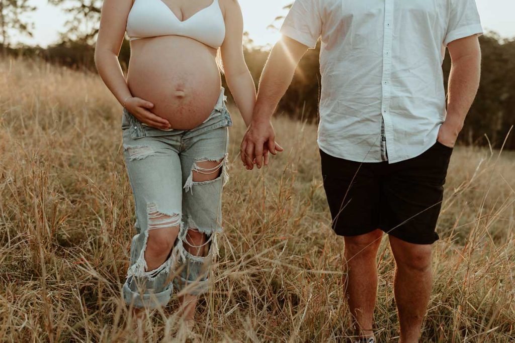 pregnant couple holding hands in a field with long grass at golden hour
