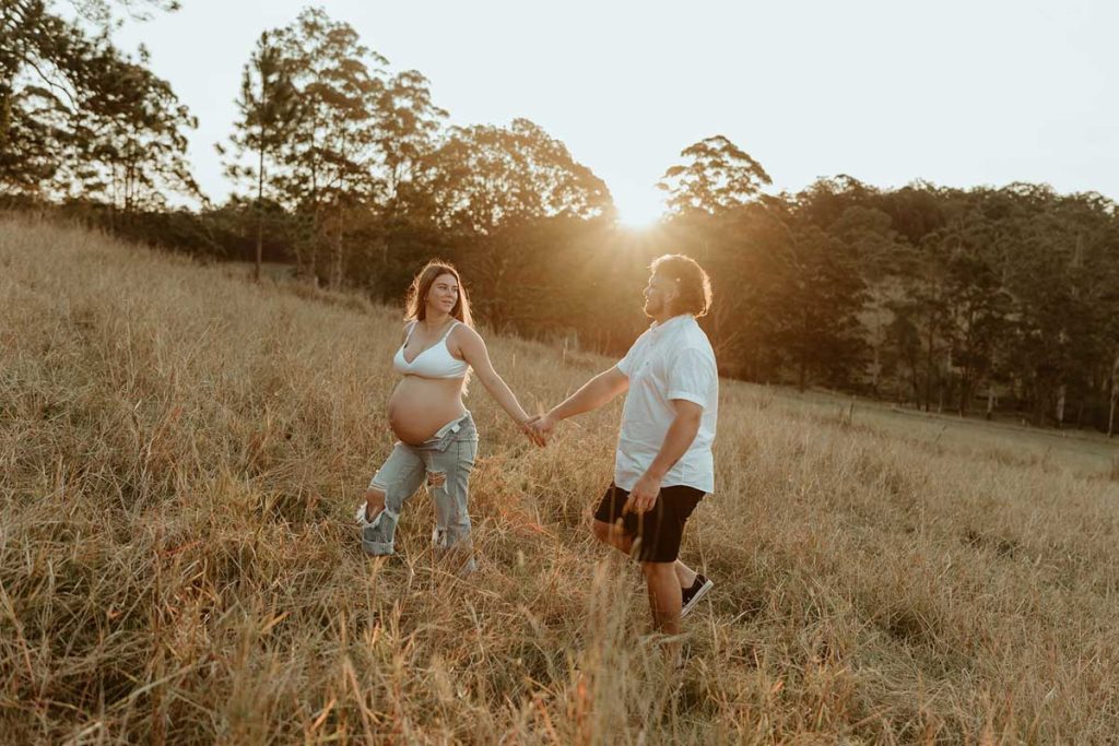 pregnant couple walking in a field with long grass looking at each other smiling at golden hour