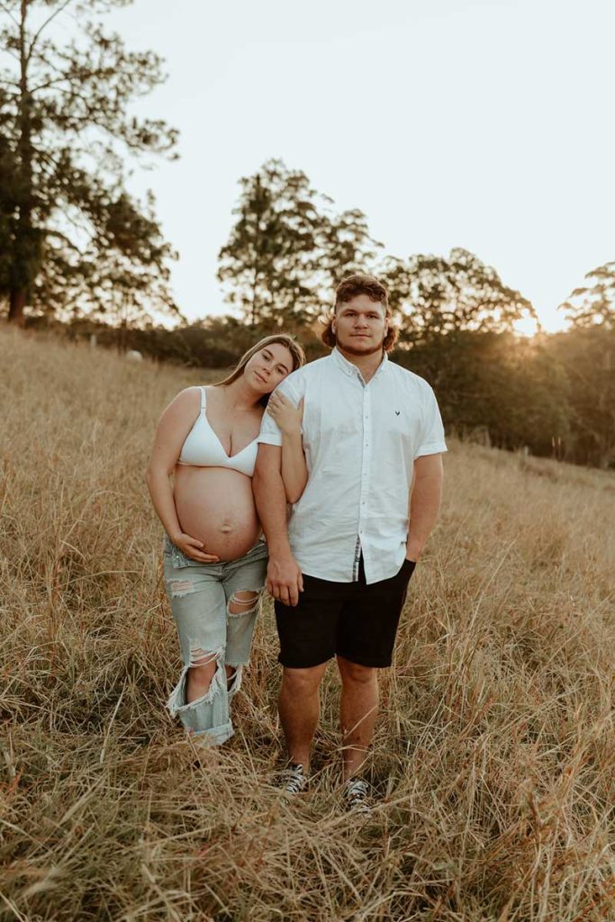 pregnant couple standing next to one another in a field with long grass