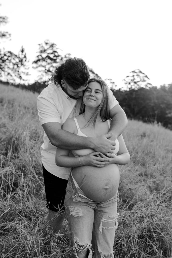 black and white image of a pregnant couple cuddling standing up in a field