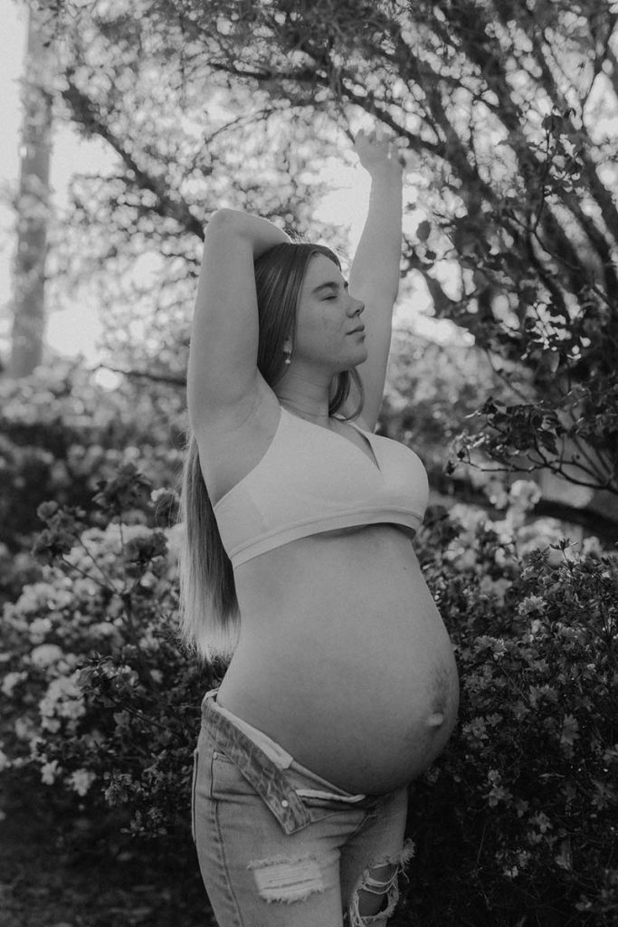 black and white image of a pregnant young woman standing near a bush of flowers