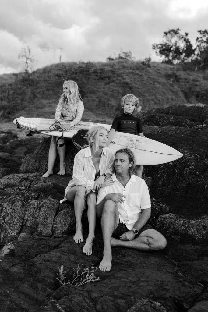 black and white image of a family sitting on a rock checking the surf and mum and dad being affectionate to each other