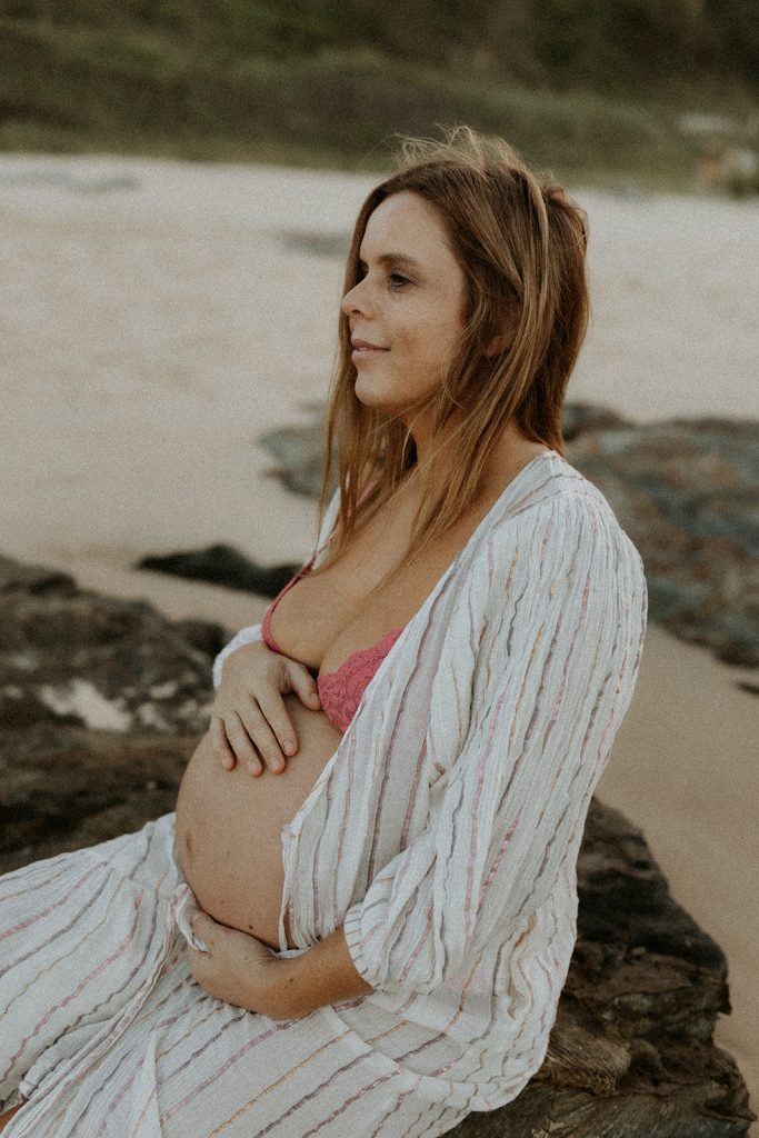 Pregnant woman sitting on a rock at the beach holding her belly and looking away